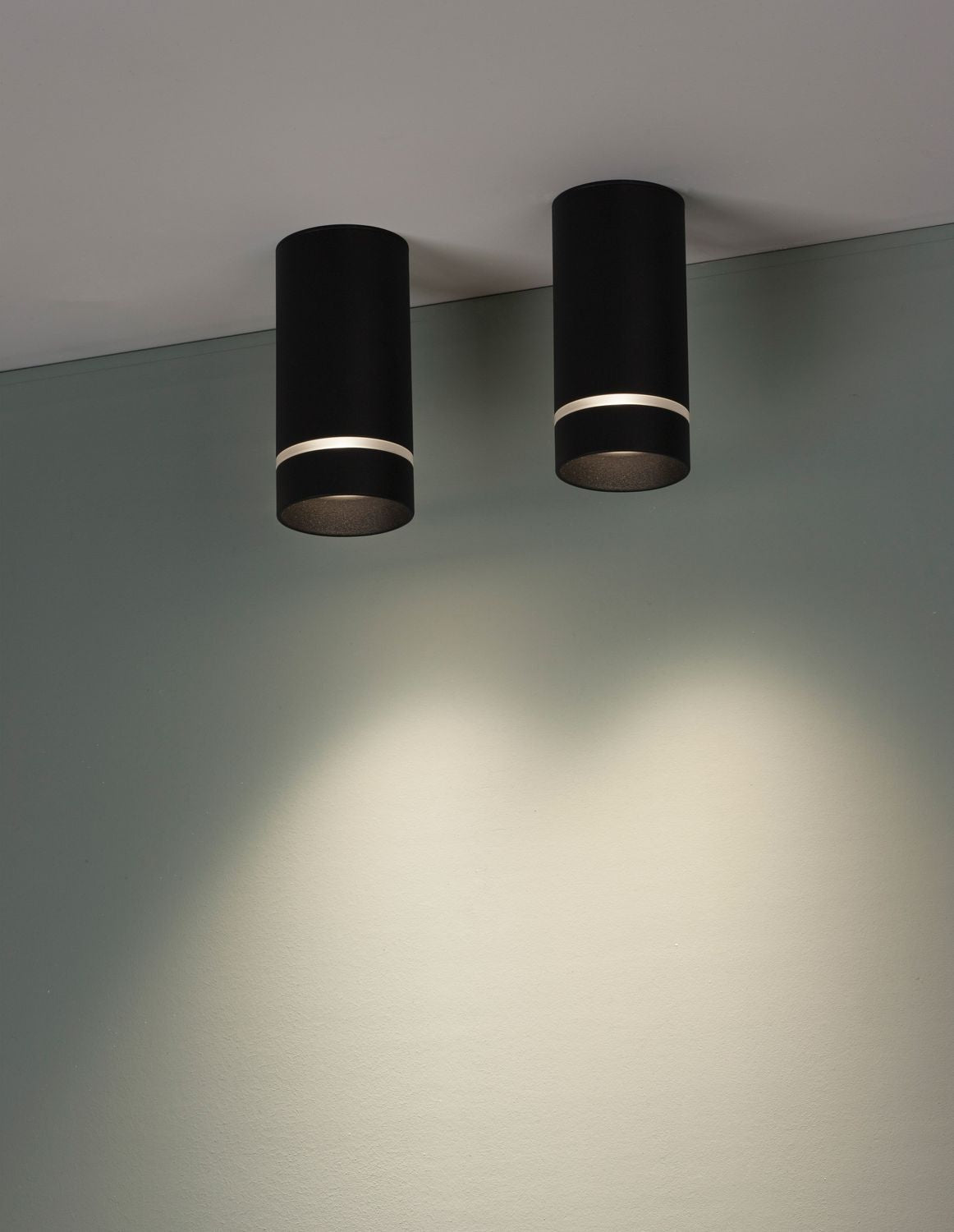 Deux plafonniers cylindriques noirs fixés au plafond éclairent un mur vert pâle.