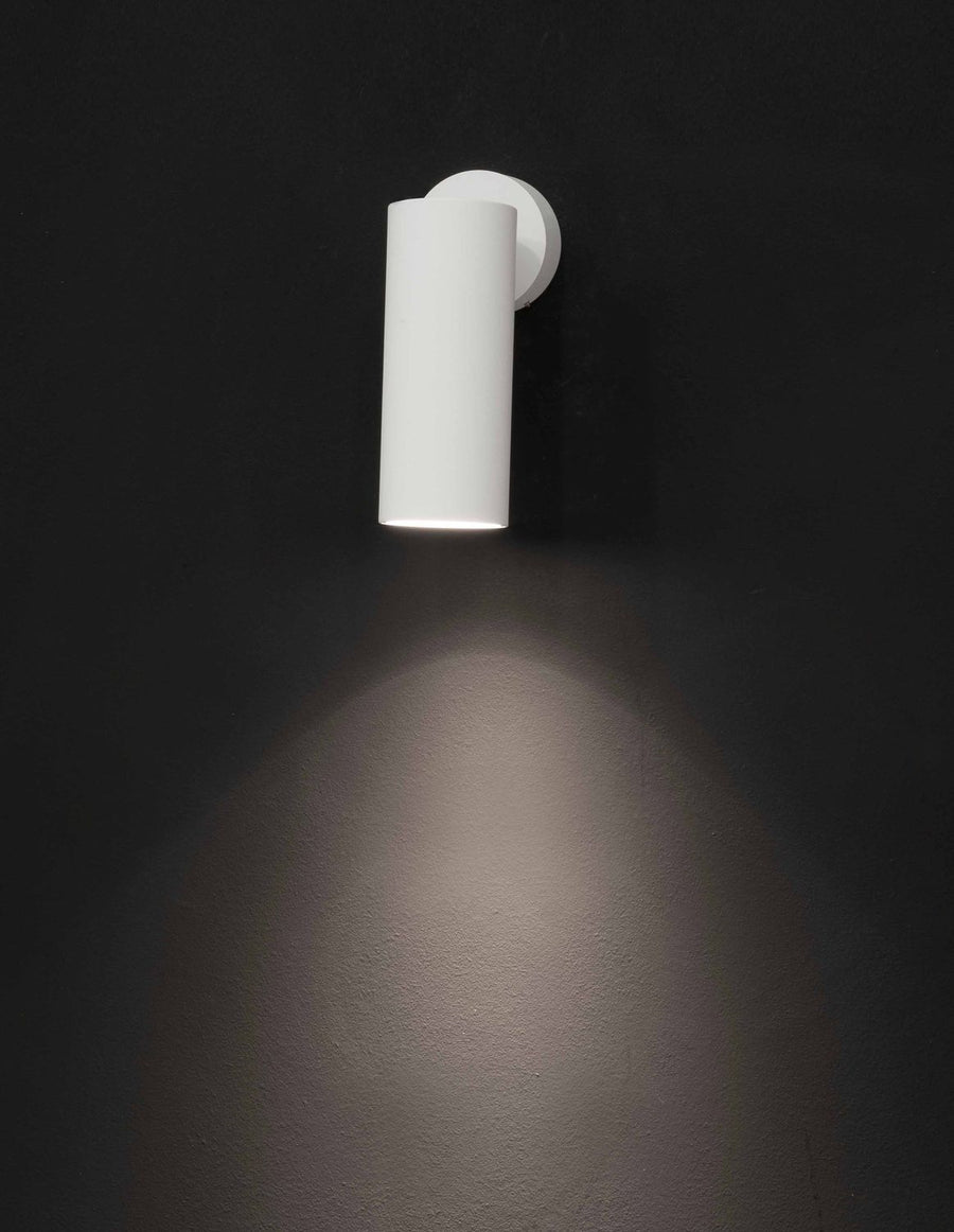 Applique murale blanche cylindrique allumée, projetant un halo lumineux sur un mur sombre.
