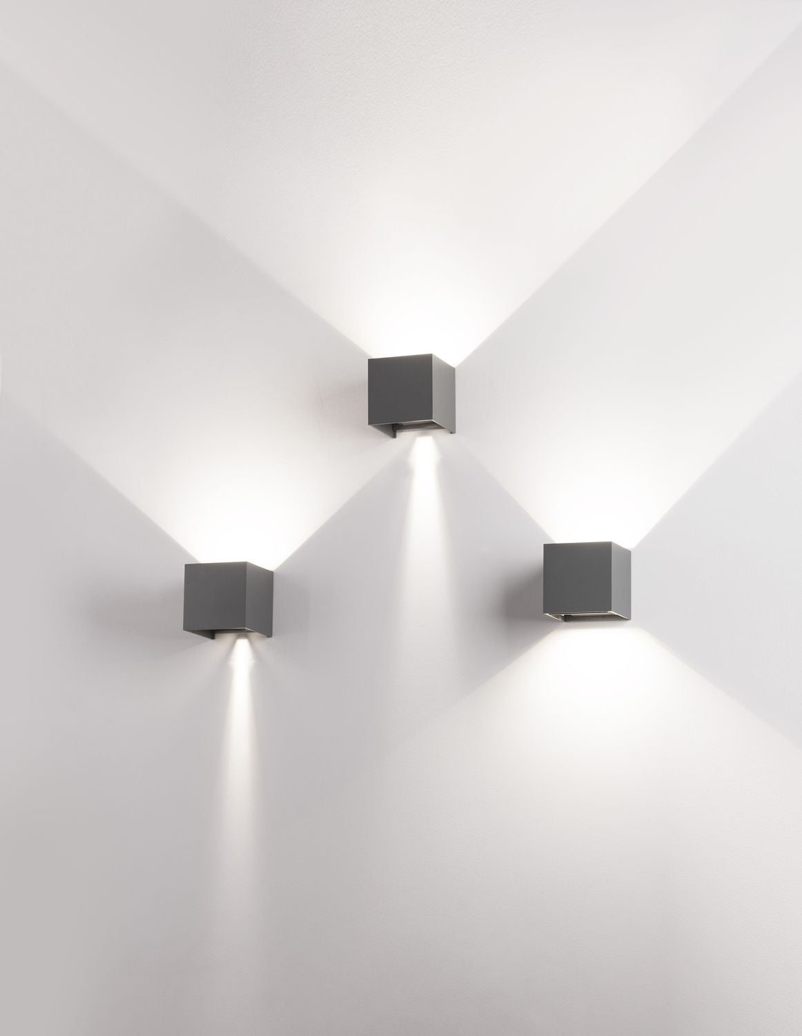 Trois appliques cubes grises fixées au mur blanc, éclairant vers le haut et le bas.