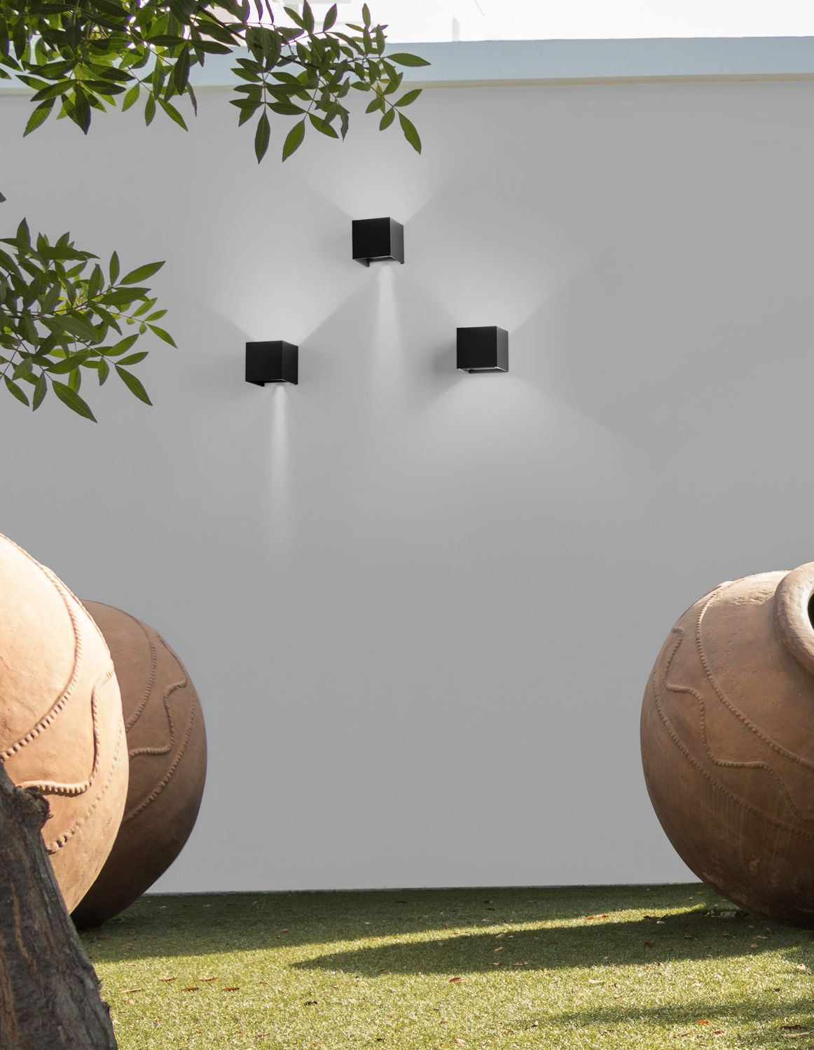 Trois appliques cubiques noires éclairant un mur blanc de jardin, ambiance extérieure avec jarres