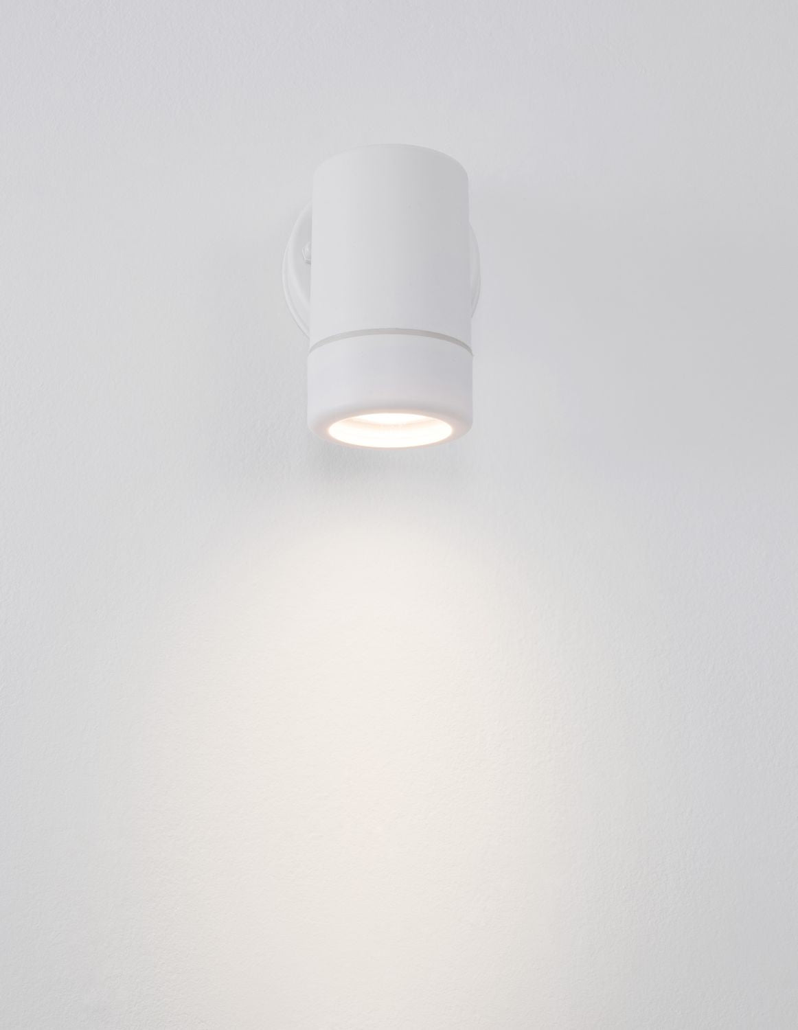 Applique cylindrique blanche allumée sur mur blanc, éclairage diffus vers le bas