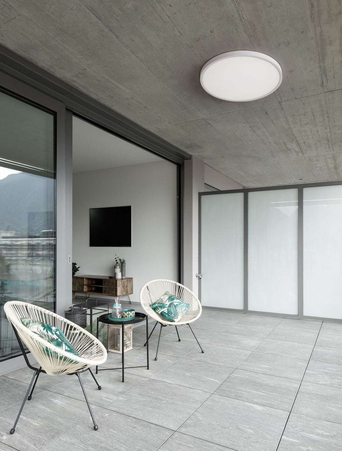 Plafonnier rond blanc fixé au plafond en béton d'une terrasse meublée de fauteuils en rotin