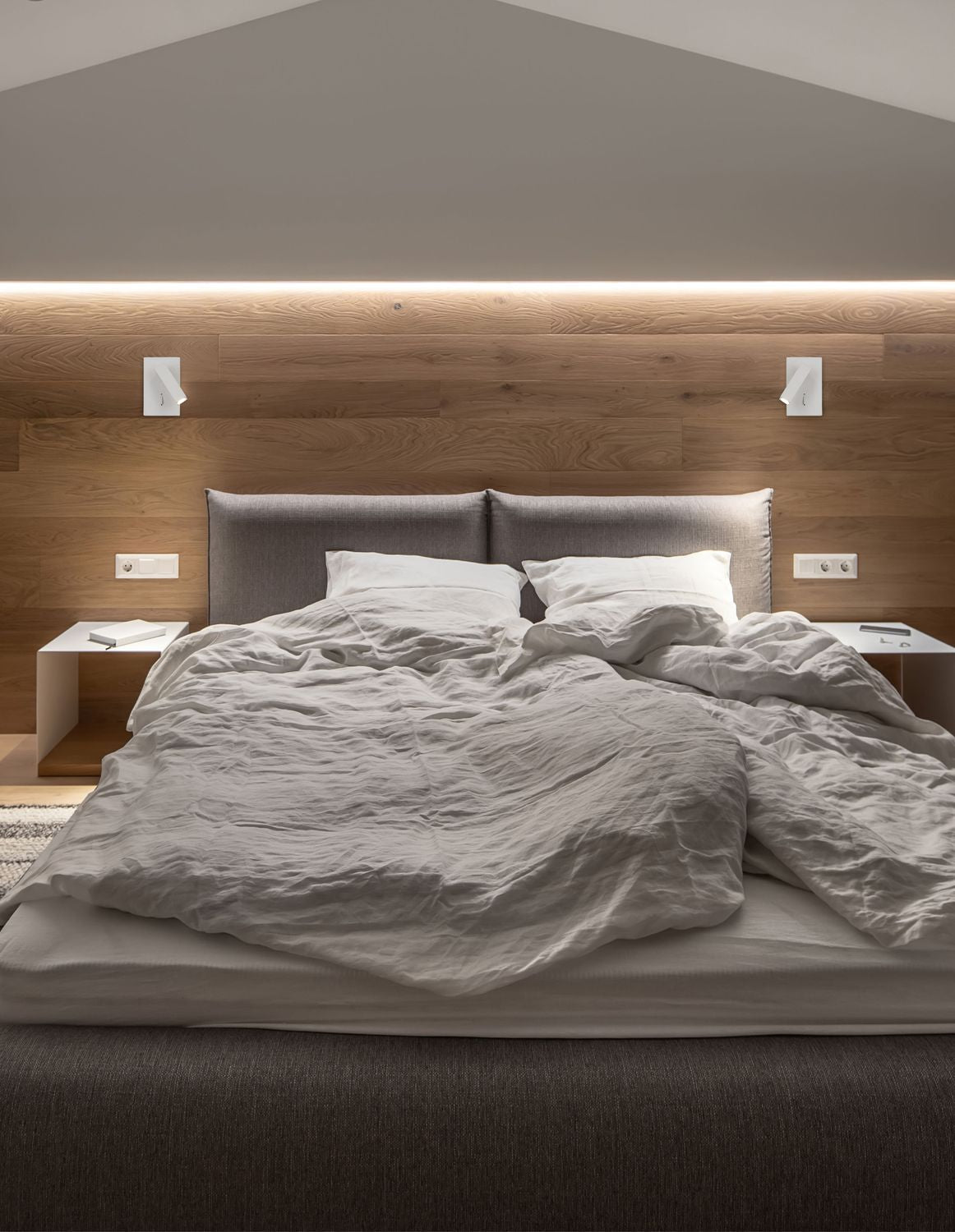 Applique murale blanche allumée au-dessus d’un lit, tête de lit en bois clair, ambiance chaleureuse.