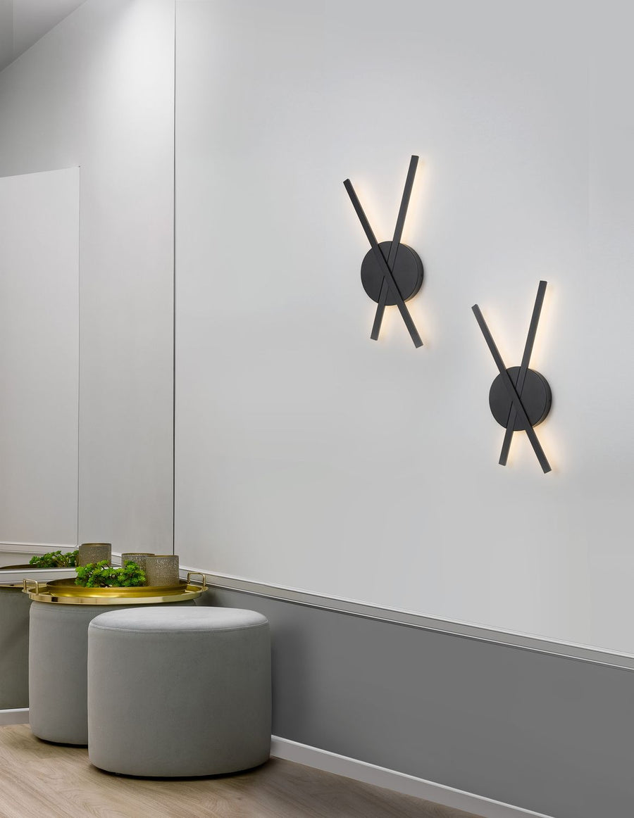 Deux appliques noires allumées sur un mur gris, au-dessus de poufs en velours