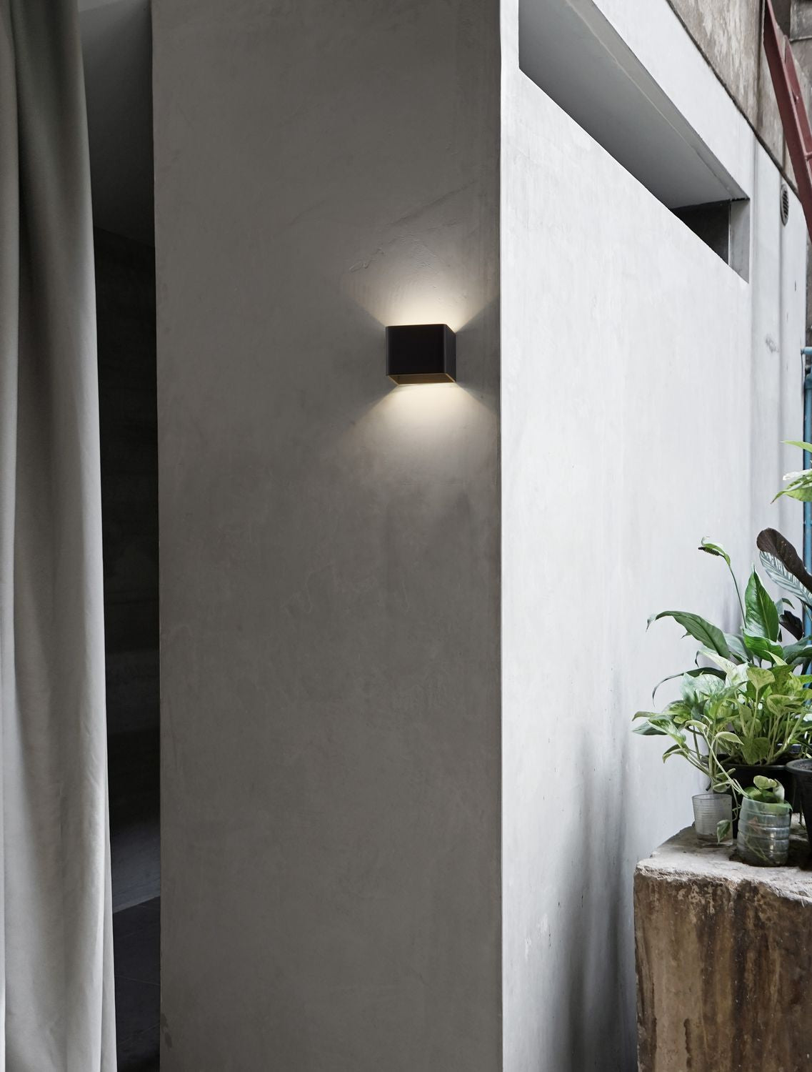 Applique noire fixée sur un mur brut, entourée d'une plante verte et d'un rideau