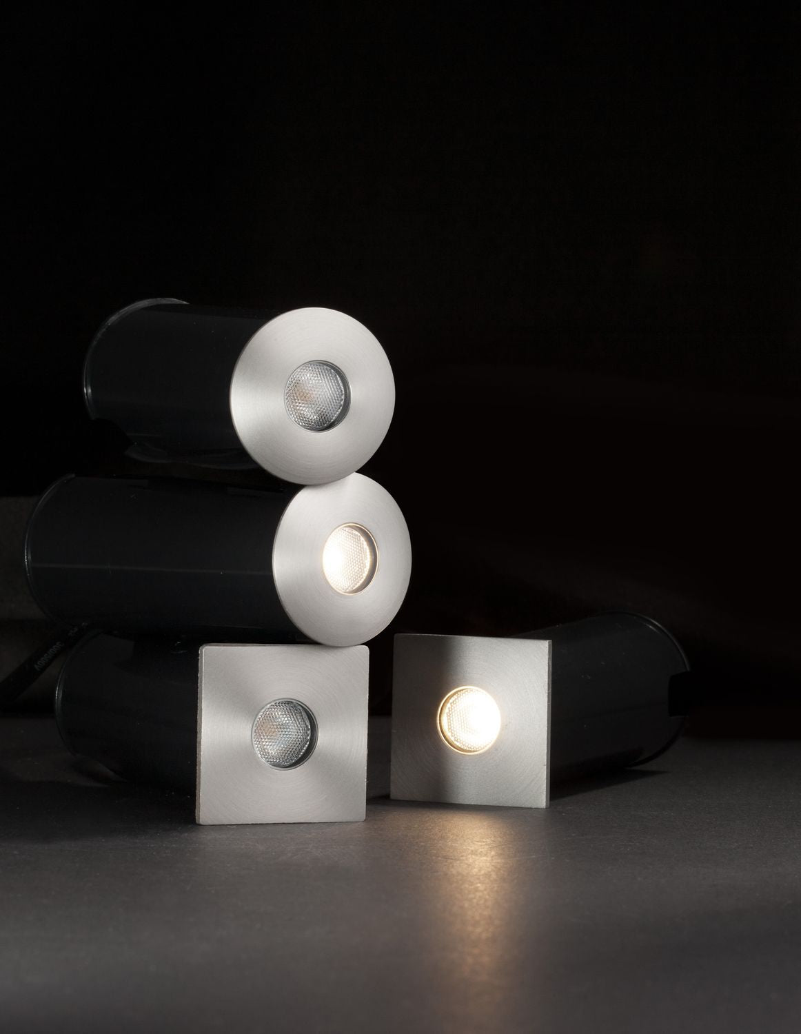 Plusieurs luminaires encastrés noirs et nickel allumés, posés sur surface sombre en ambiance nocturne.