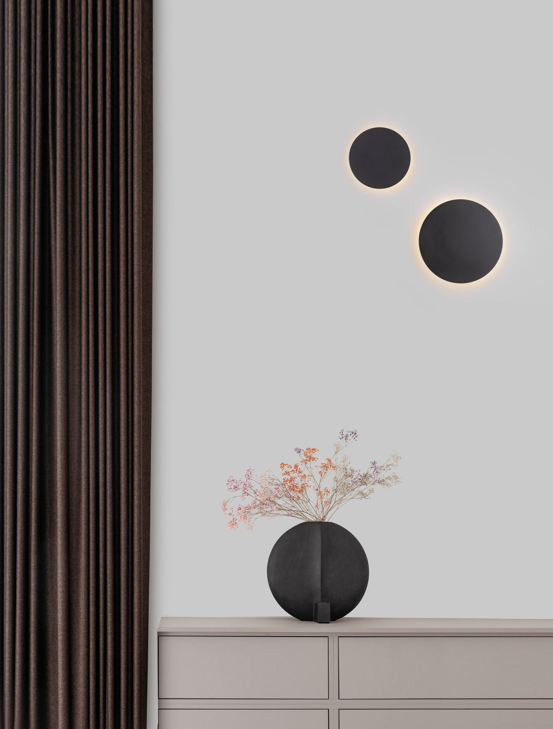 Deux appliques murales noires et rondes éclairées sur un mur, au-dessus d'un meuble et d'un vase.