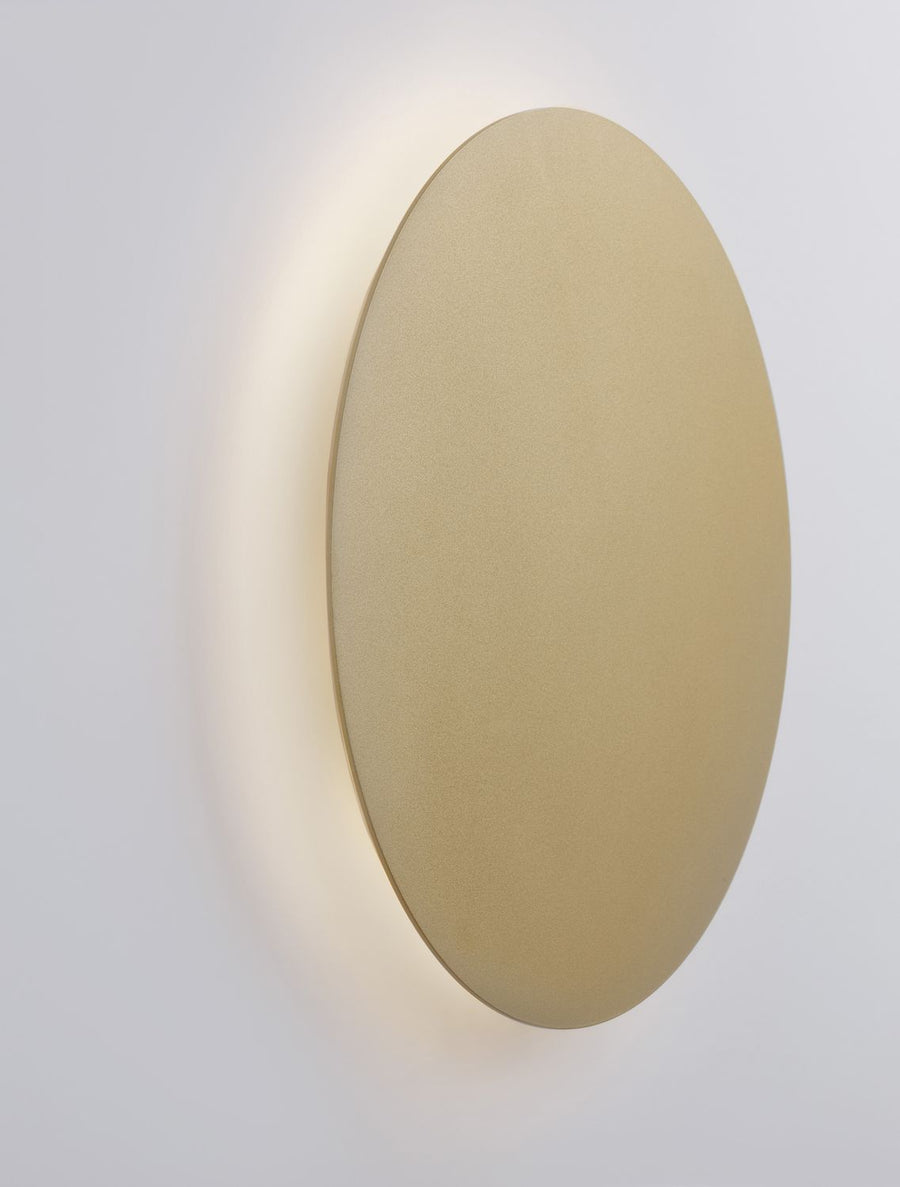 Applique murale ronde dorée vue de trois quarts, halo lumineux diffus sur le mur