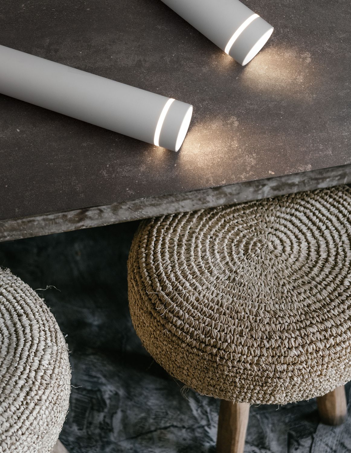 Deux suspensions cylindriques blanches posées sur une table sombre, ambiance chaleureuse avec poufs en jute