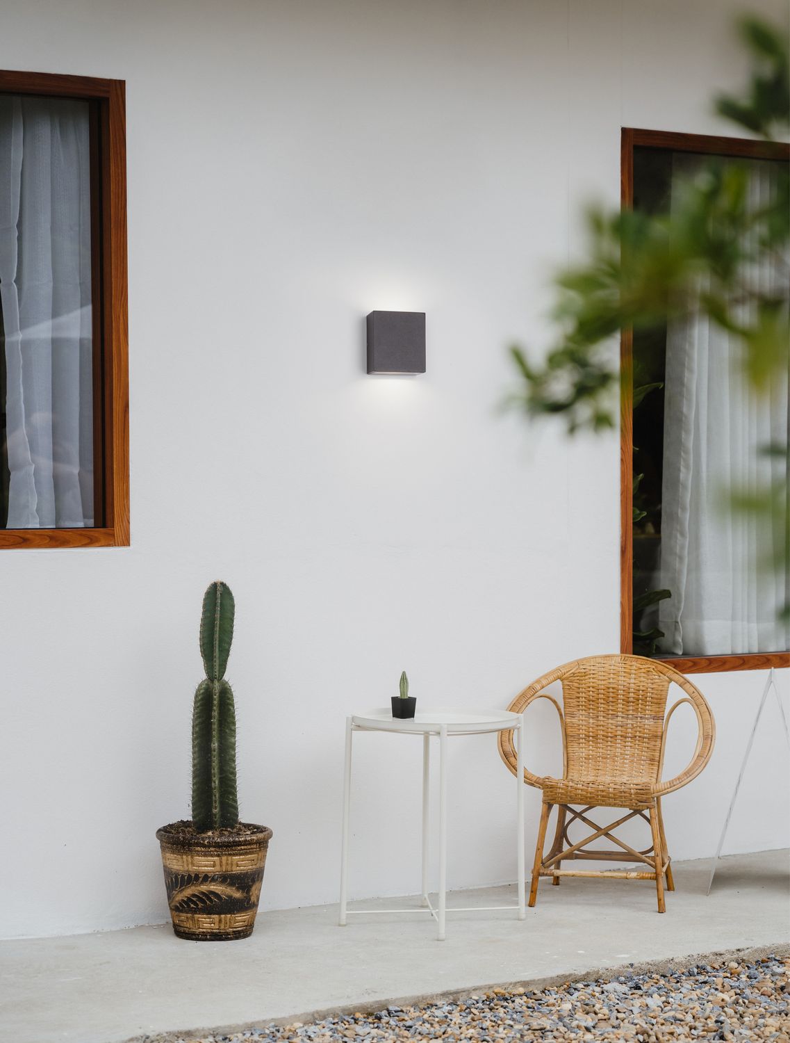 Applique murale anthracite allumée sur mur blanc, terrasse et fauteuil en rotin