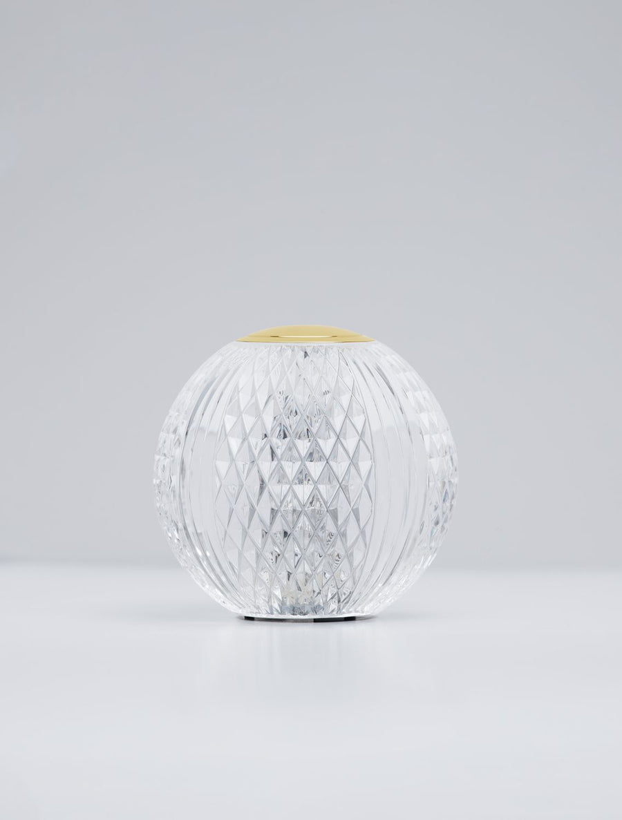 Lampe à poser sphérique en verre transparent facetté, capuchon doré, sur fond clair.