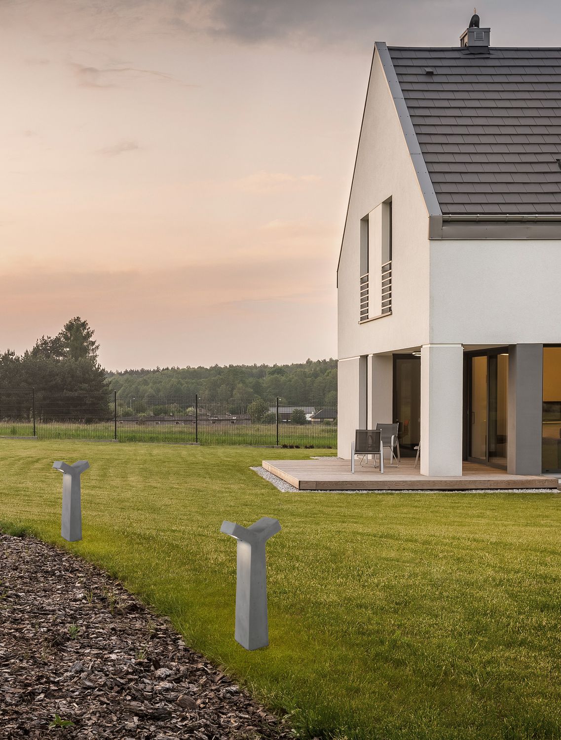 Borne extérieure grise installée dans une pelouse, mise en ambiance au crépuscule devant une maison.