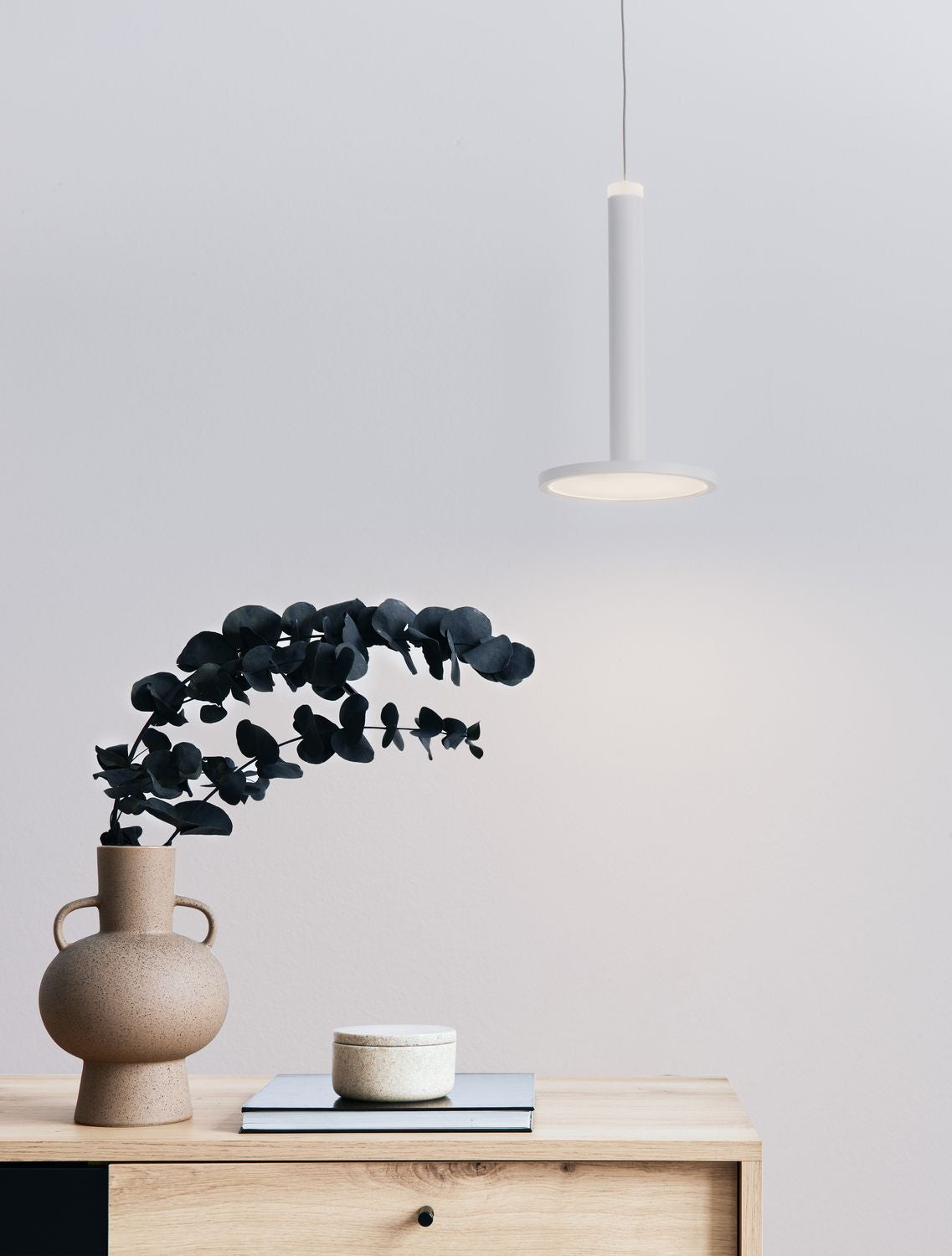 Suspension blanche allumée au-dessus d'un meuble en bois clair avec vase et eucalyptus