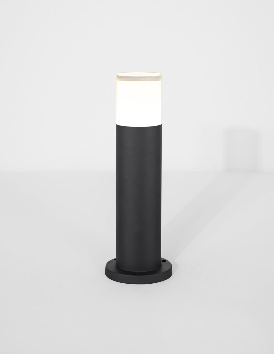 Luminaire allumé de face sur fond neutre, halo blanc au sommet du fût noir.
