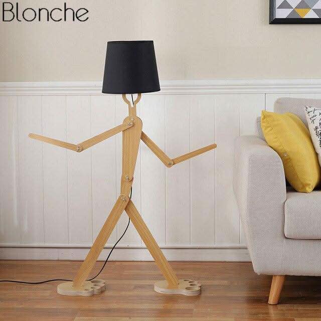 Lampadaire personnage articulé avec abat-jour noir, posé près canapé beige et coussin.