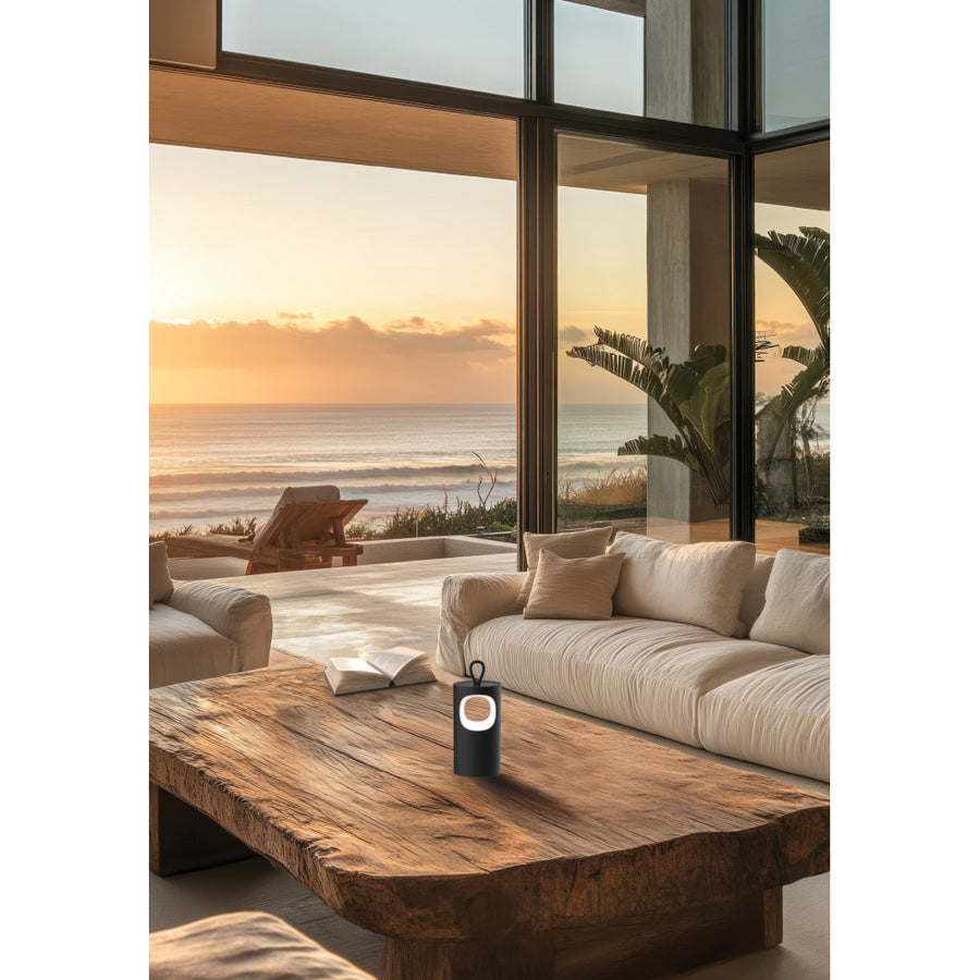 Lampe noire posée sur table basse en bois devant canapé, vue mer au coucher du soleil.