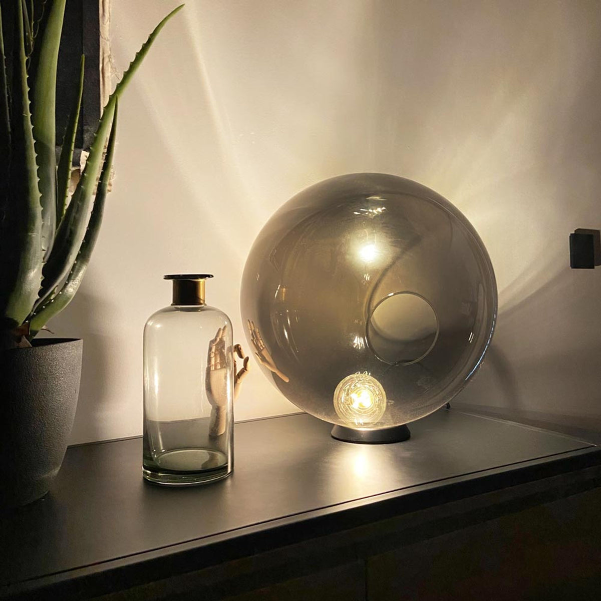 Un globe en verre fumé sur un meuble noir voisinant un vase et un aloès