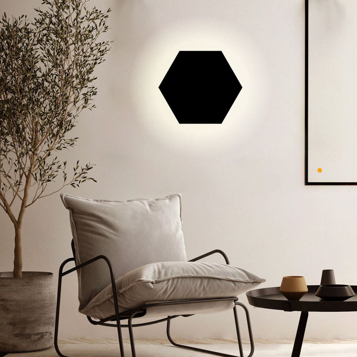 Version noire de l'applique hexagonale murale, allumée, dans un salon avec fauteuil et plante.