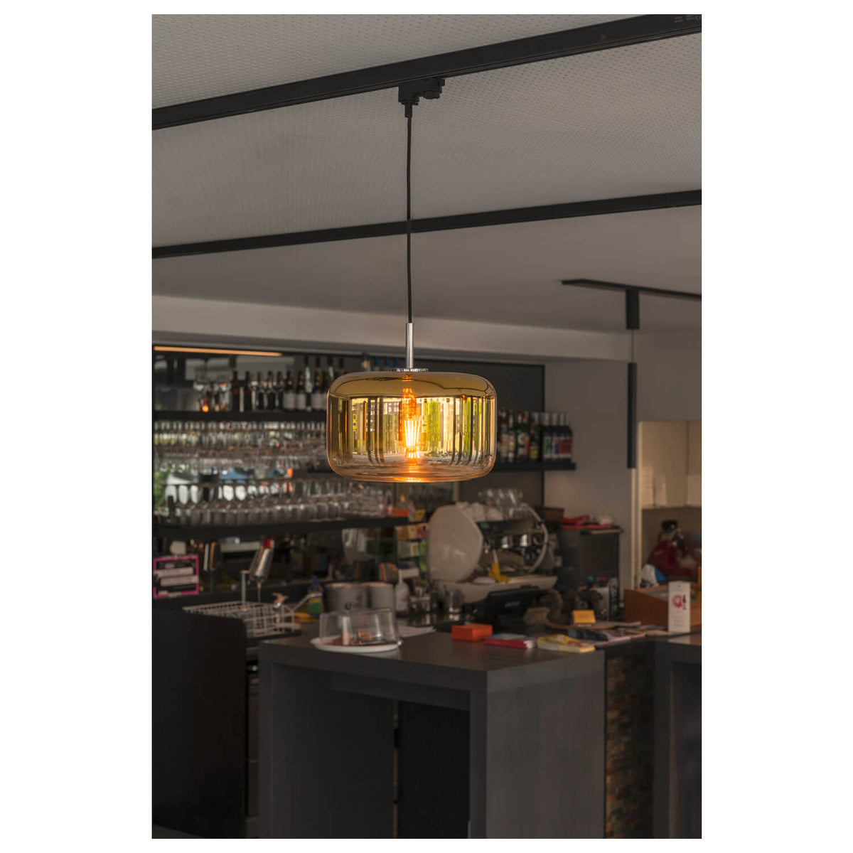 Mise en ambiance au-dessus d’un comptoir de bar, suspension dorée et bouteilles en arrière-plan.