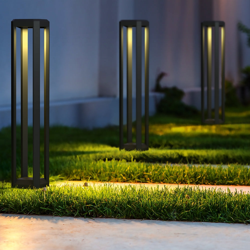 Mise en ambiance nocturne dans un jardin : trois bornes grises éclairent gazon et allée.