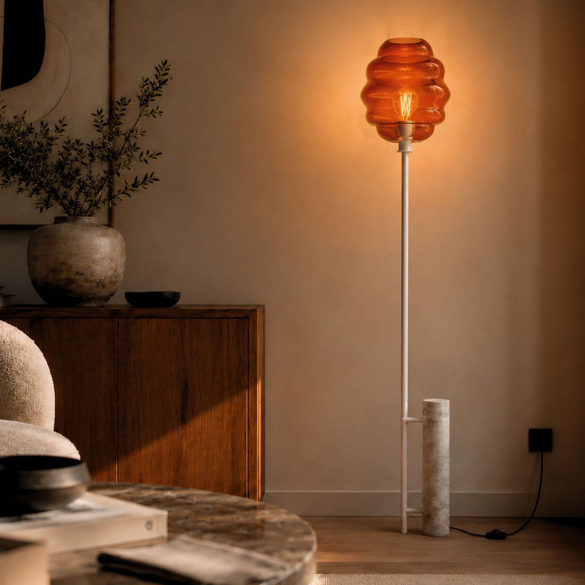 Lampadaire allumé dans un intérieur, abat-jour en verre ambré et pied travertin visible.