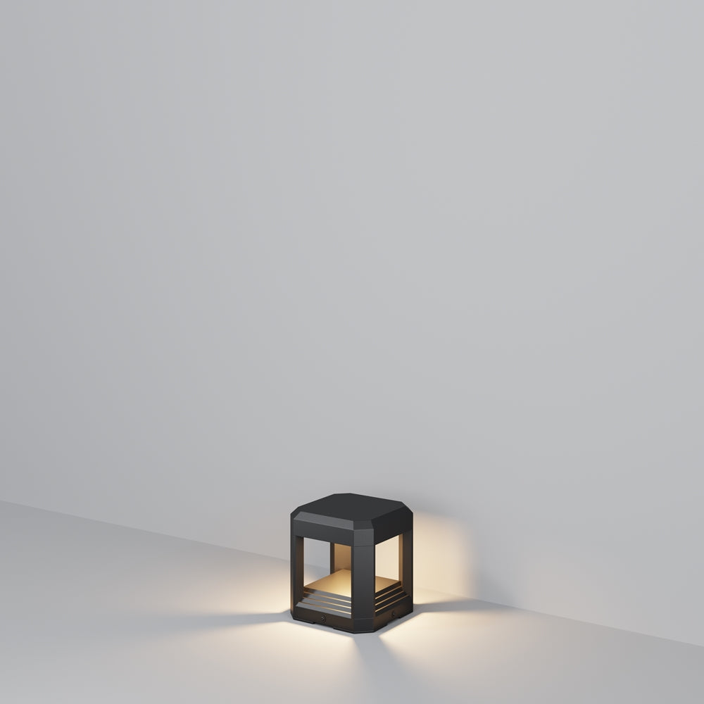 Petit luminaire cubique gris foncé posé au sol, lumière chaude diffusée sous la base.
