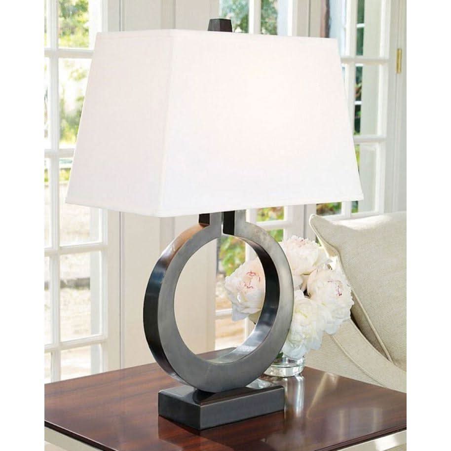 Lampe sur table devant une fenêtre, base métallique foncée et abat-jour blanc, fleurs à l'arrière-plan.