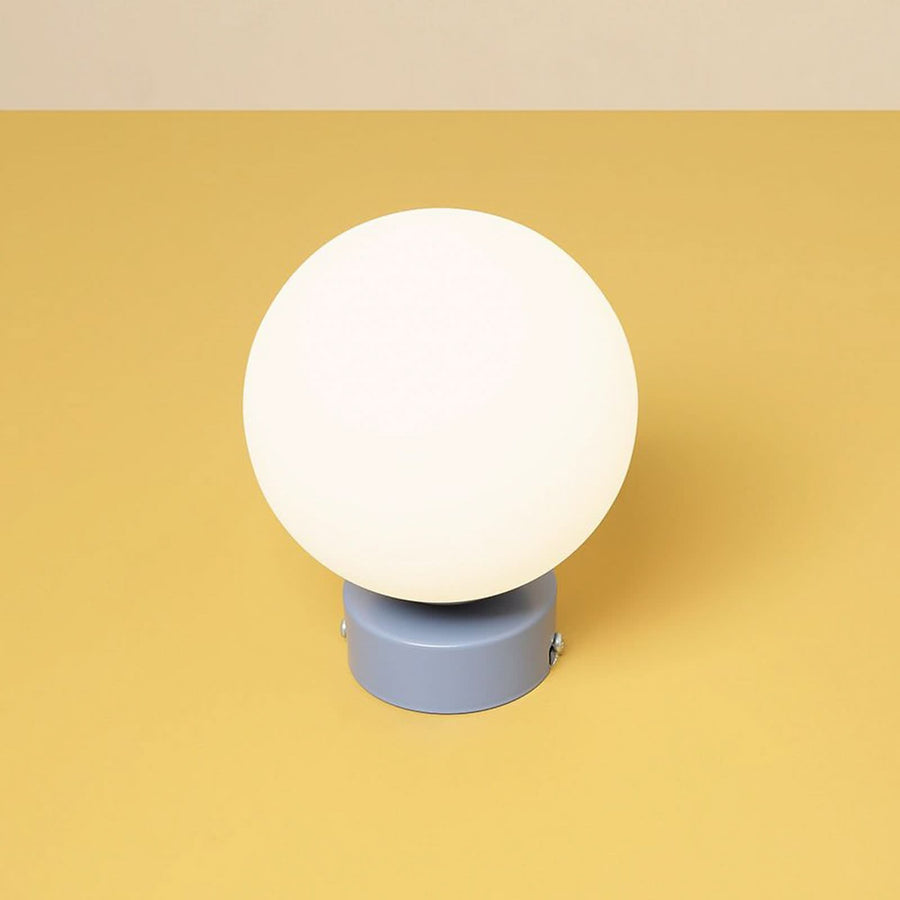 Luminaire présenté de face sur fond jaune, sphère blanche sur base cylindrique.