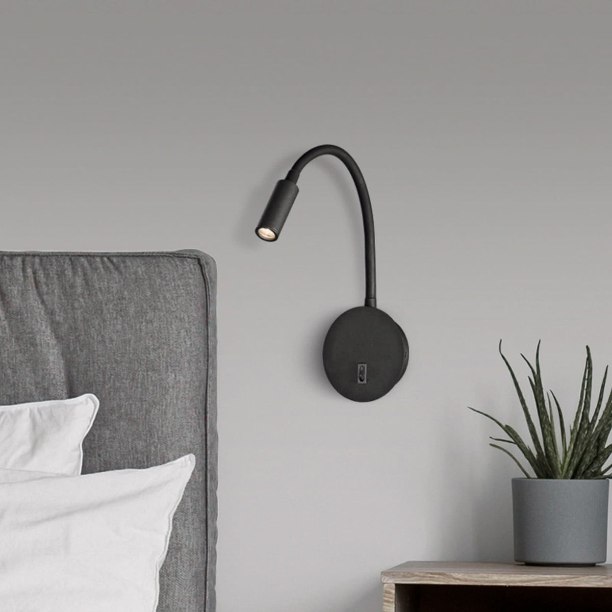 Applique murale noire flexible installée près d'une tête de lit grise avec plante.