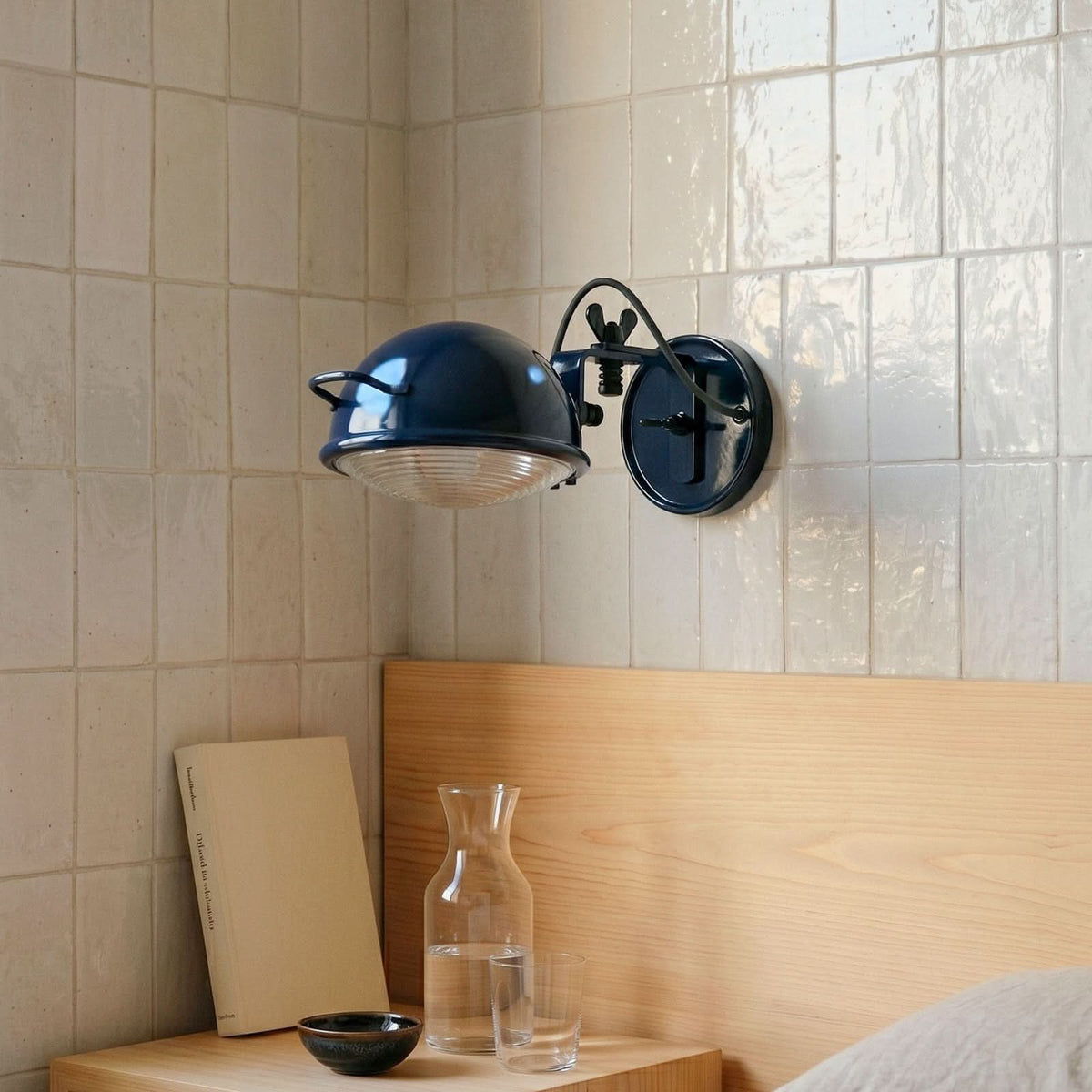 Luminaire mural bleu installé dans une salle de bain carrelée, au-dessus d’un chevet en bois.