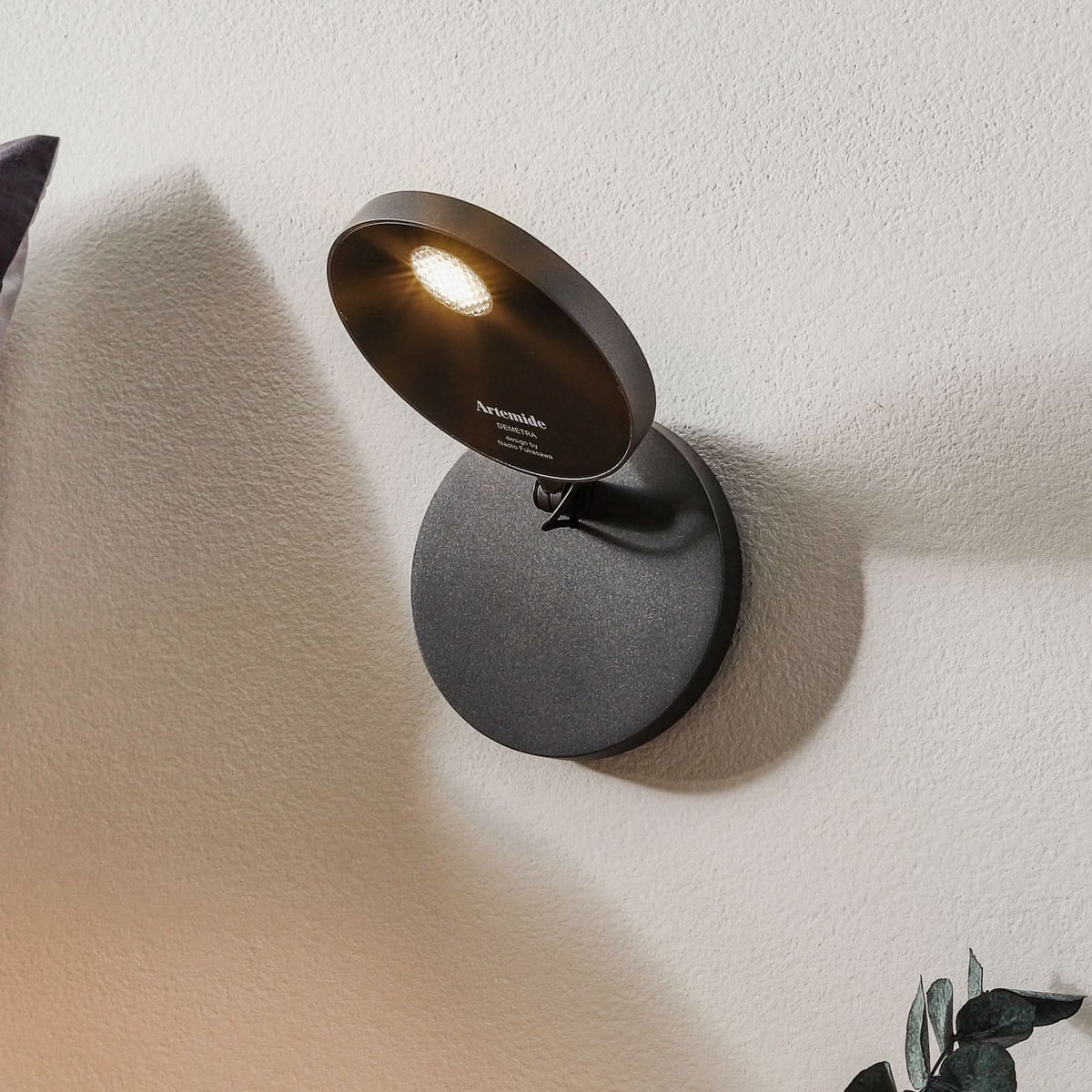 Applique murale ronde en aluminium anthracite, allumée et orientée vers le haut sur un mur.