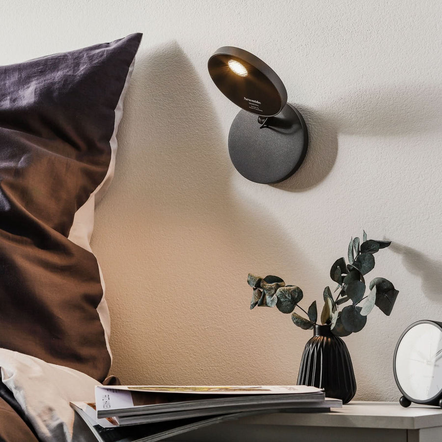 Vue en situation du luminaire fixé près d'un lit, avec coussin, magazines et plante.
