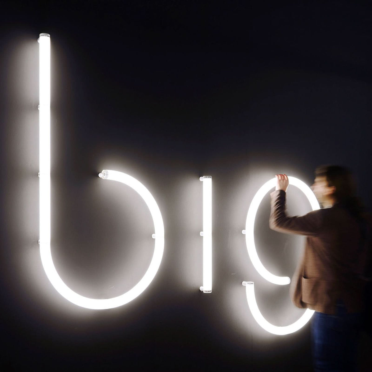 Une personne installe ou touche les lettres lumineuses big sur un mur sombre.