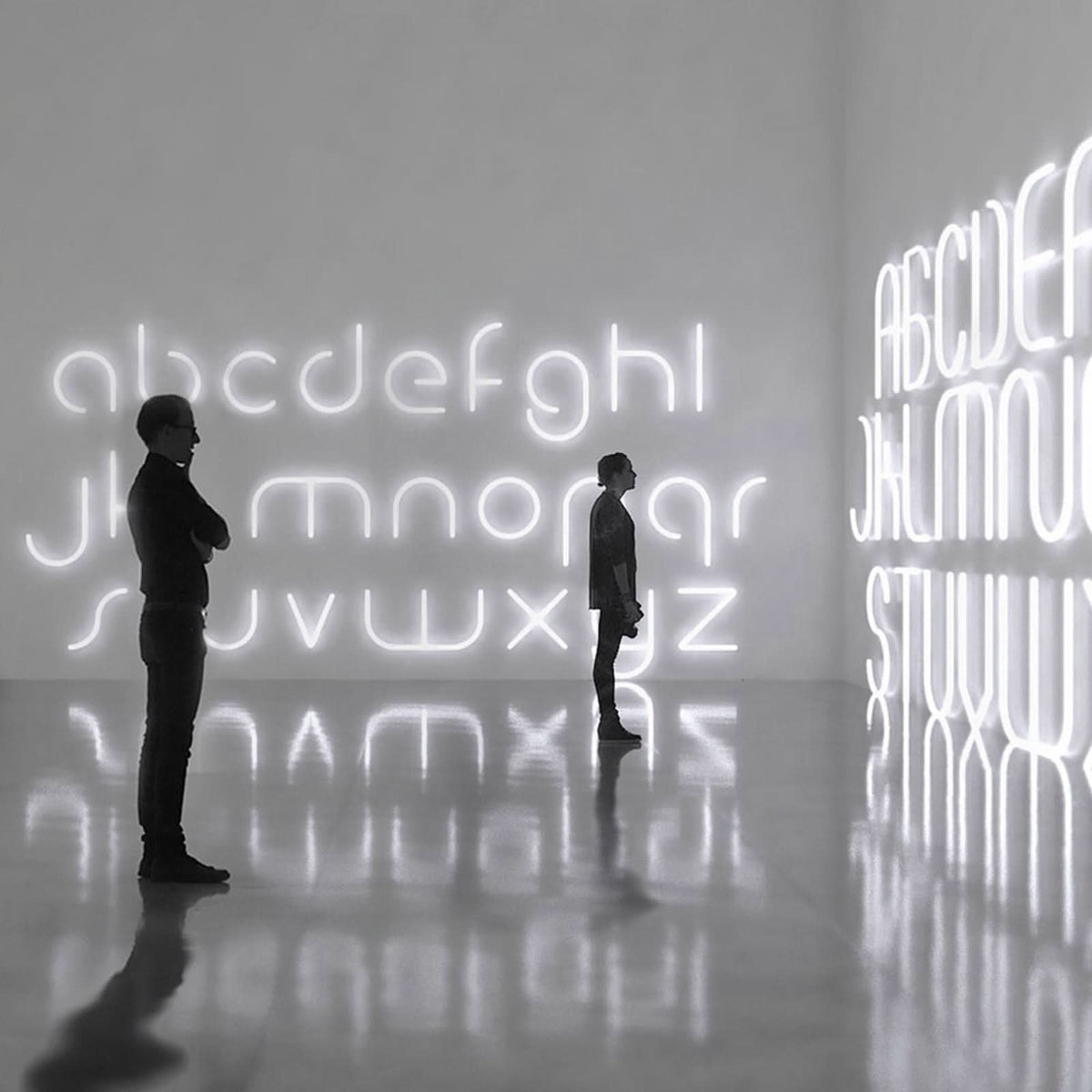 Deux personnes contemplent des lettres lumineuses géantes dans une galerie au sol réfléchissant.