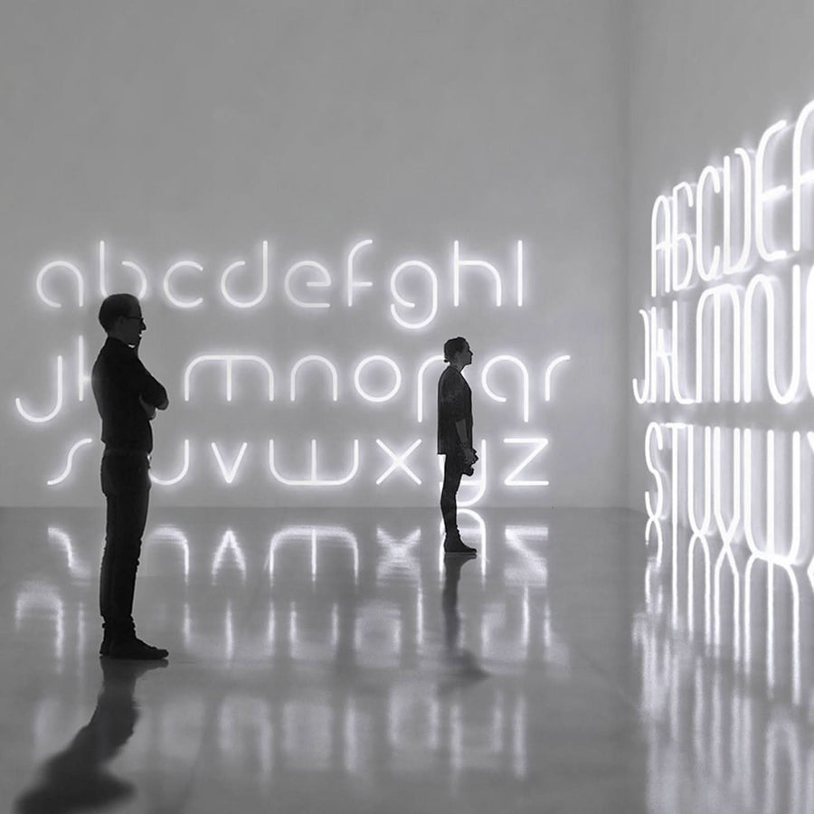 Deux silhouettes contemplent des lettres lumineuses blanches dans une galerie sombre.