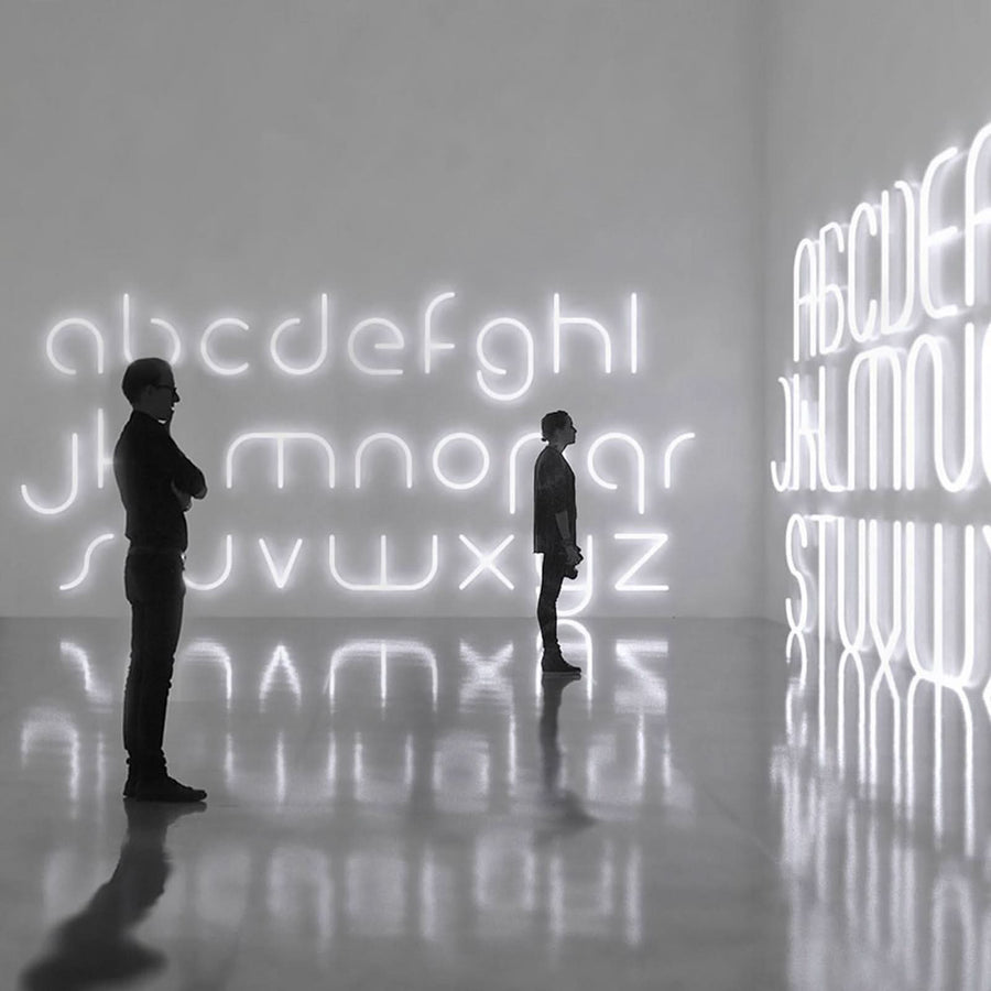 Deux silhouettes observent une grande installation murale d’alphabet lumineux dans une galerie réfléchissante.