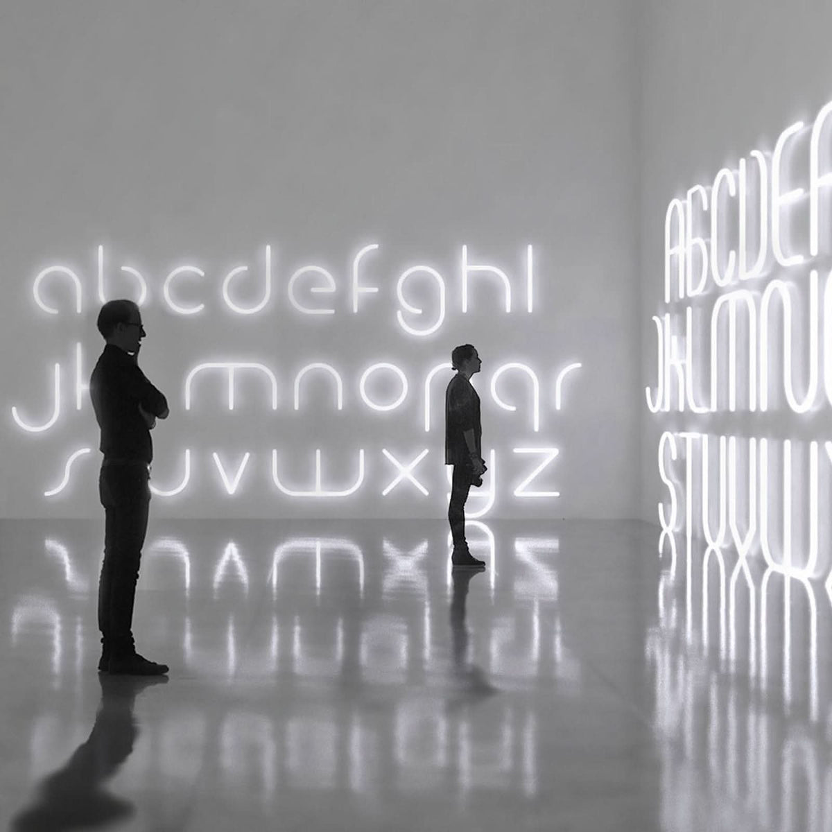 Deux personnes observent une grande installation murale de lettres lumineuses dans une galerie.
