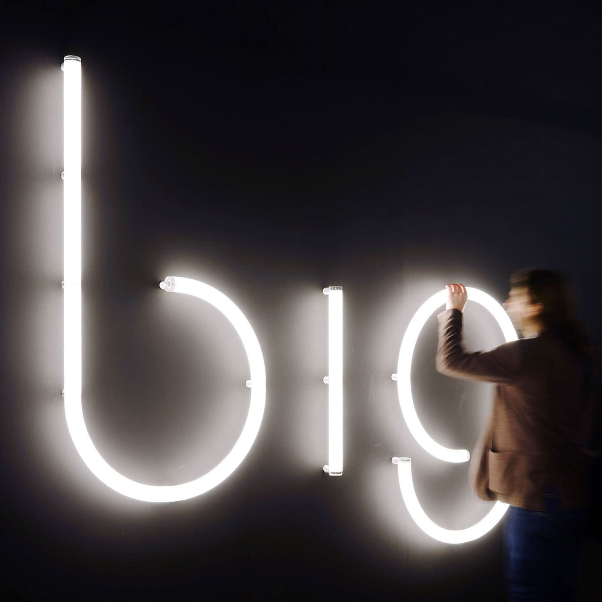 Une personne ajuste une grande lettre lumineuse blanche fixée sur un mur noir.
