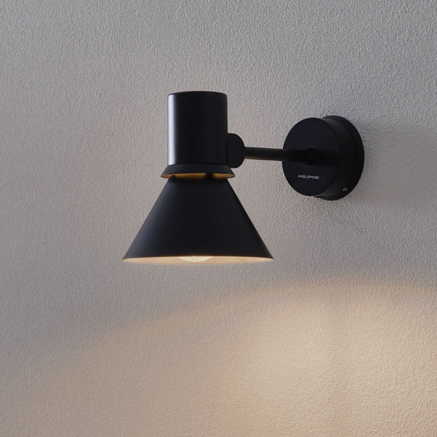 Applique murale noire fixée sur un mur texturé gris, éclairée et orientée vers le bas.