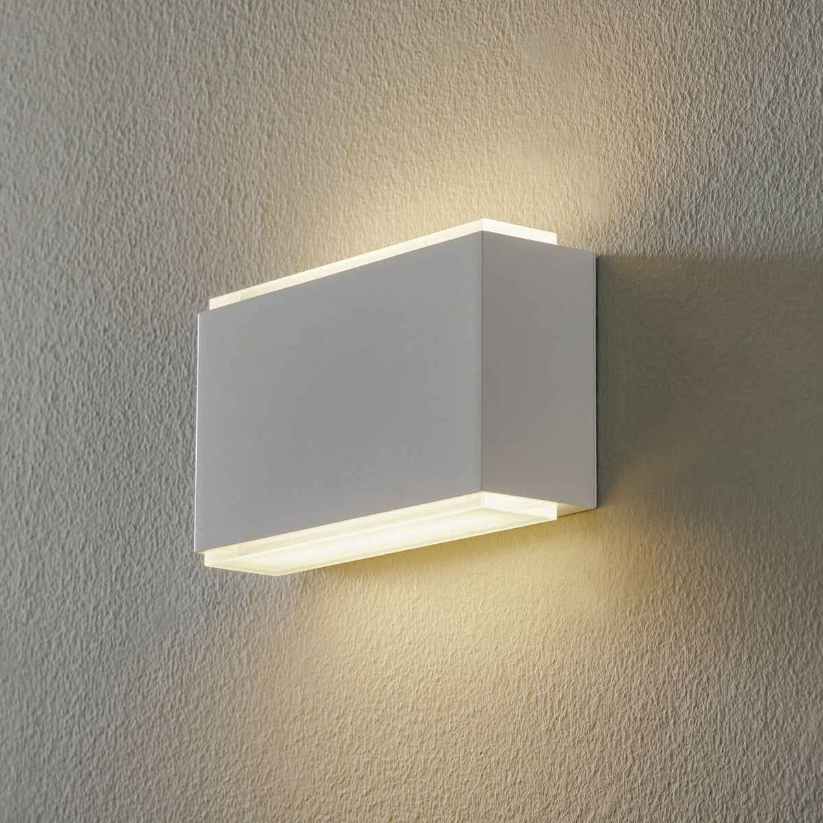 Applique murale rectangulaire blanche allumée, diffusant une lumière chaude sur un mur texturé beige.
