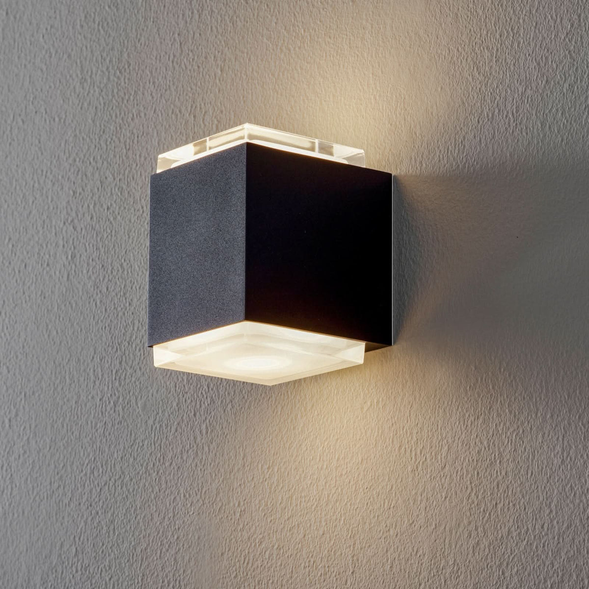 Applique murale cubique noire allumée sur un mur gris clair, lumière haut et bas