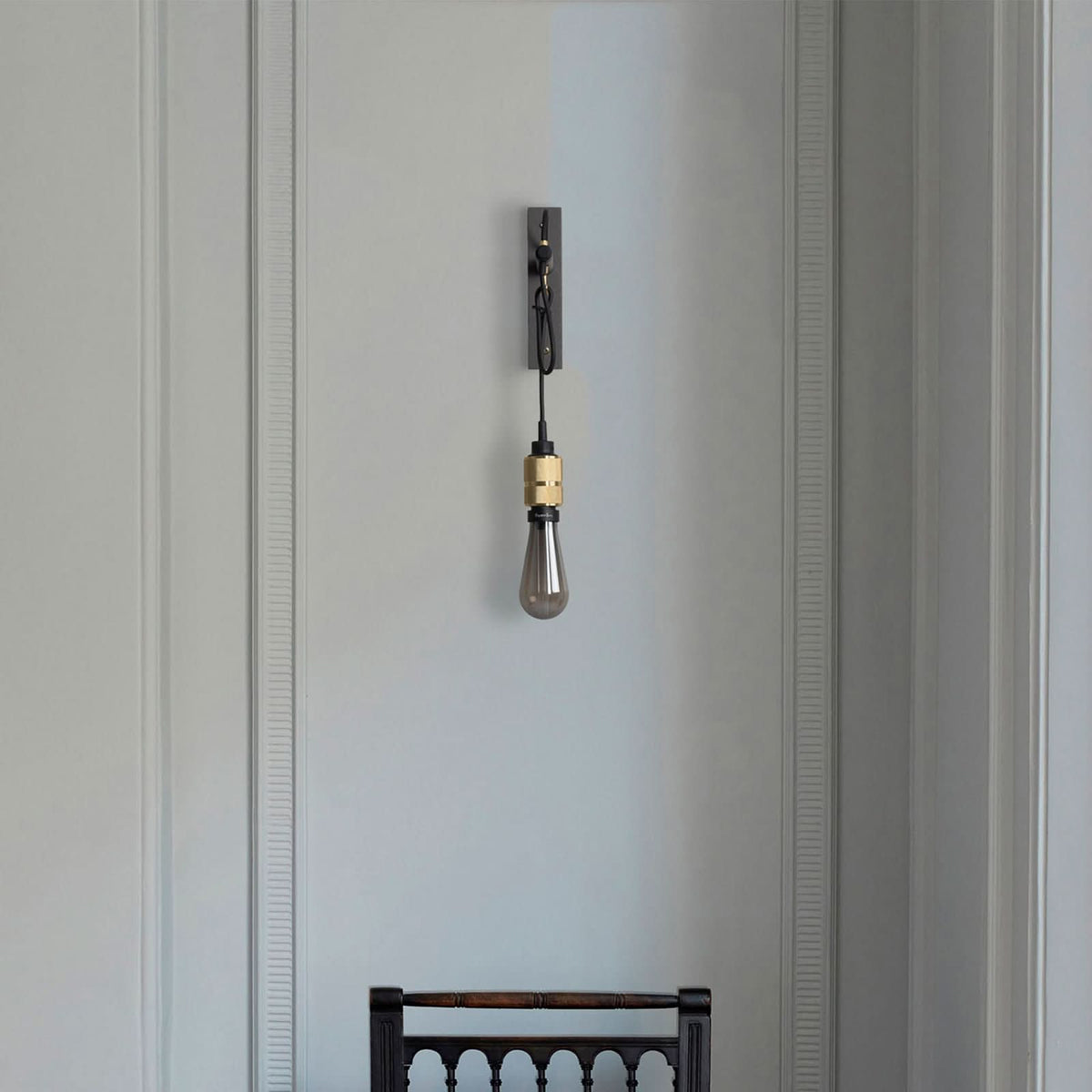Applique installée sur mur mouluré clair, ampoule éteinte au-dessus d'une chaise en bois