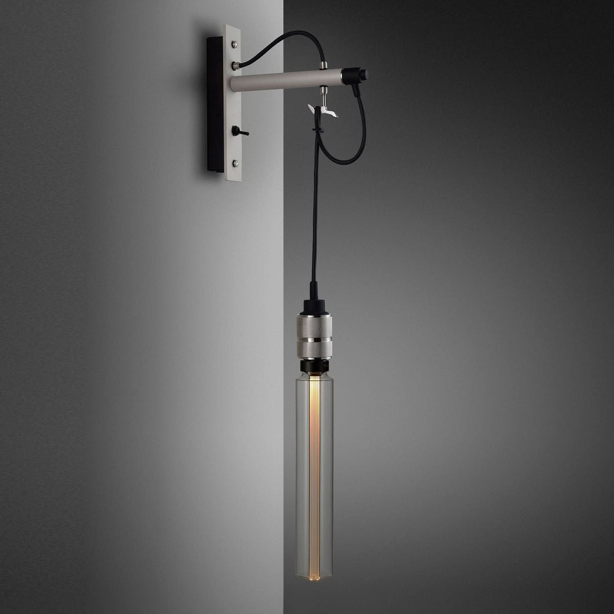 Applique murale équipée d’une longue ampoule tubulaire allumée, câble noir et support acier gris.