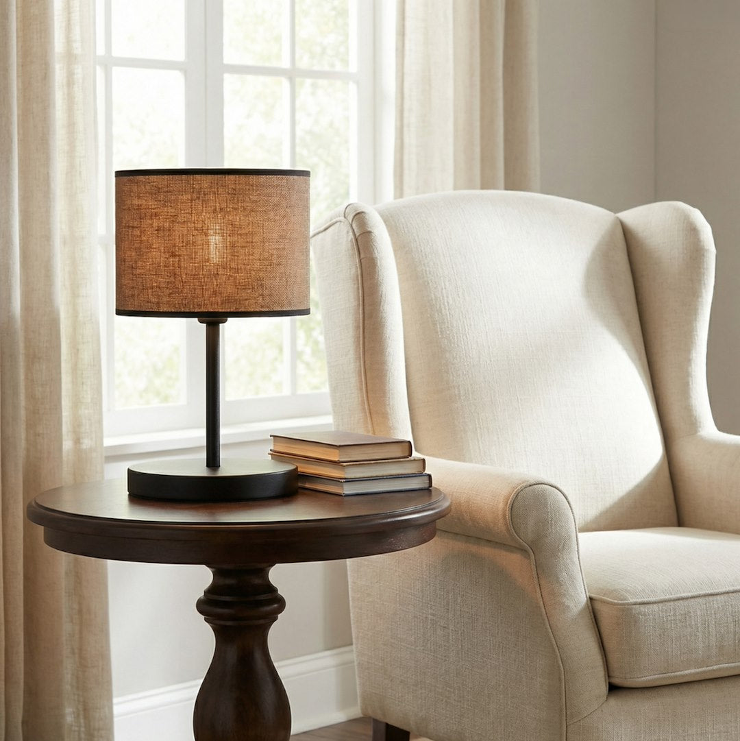 Lampe sur table ronde foncée devant une fenêtre, fauteuil crapaud beige