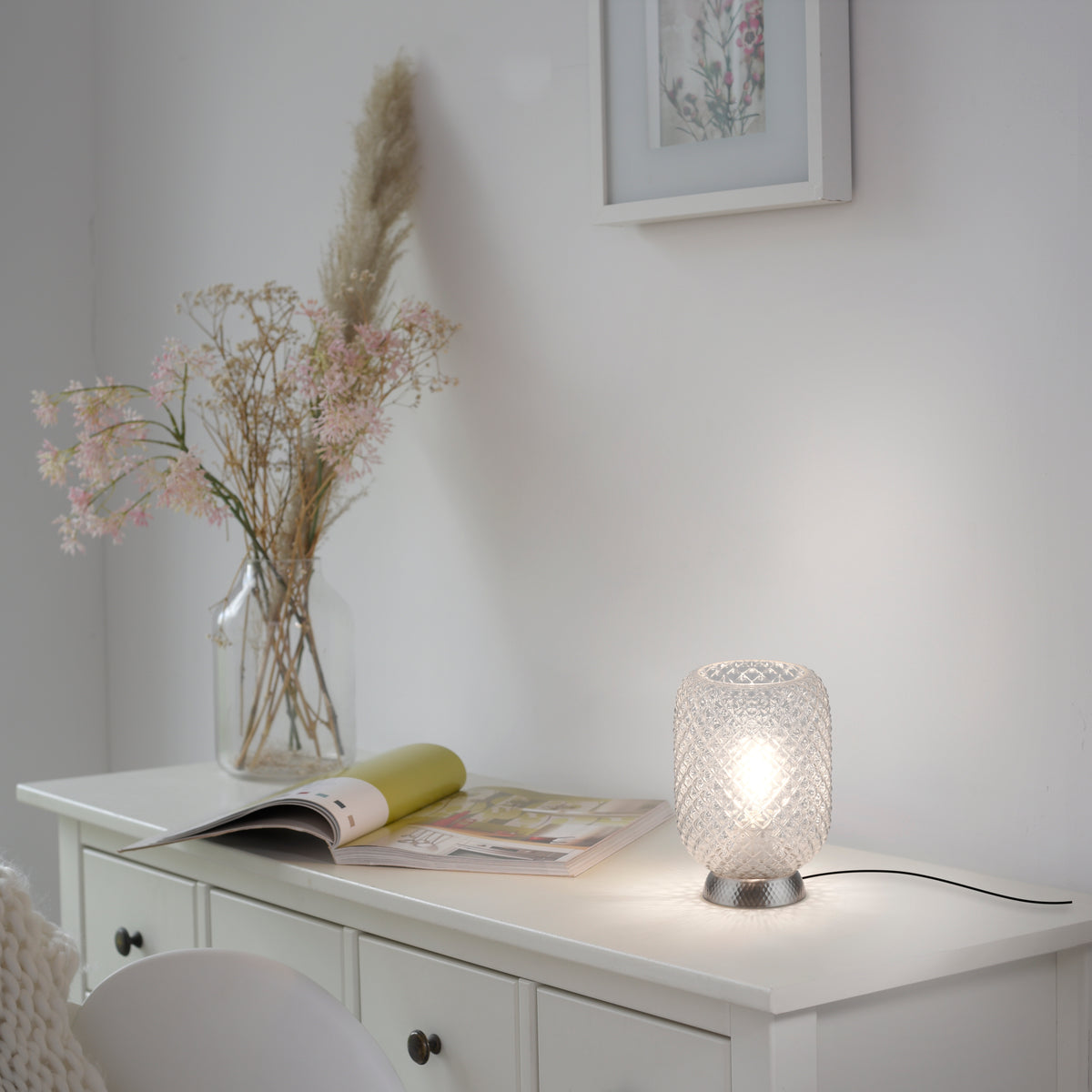 Mise en ambiance sur un meuble blanc, lampe allumée près d'un magazine et d'un vase.