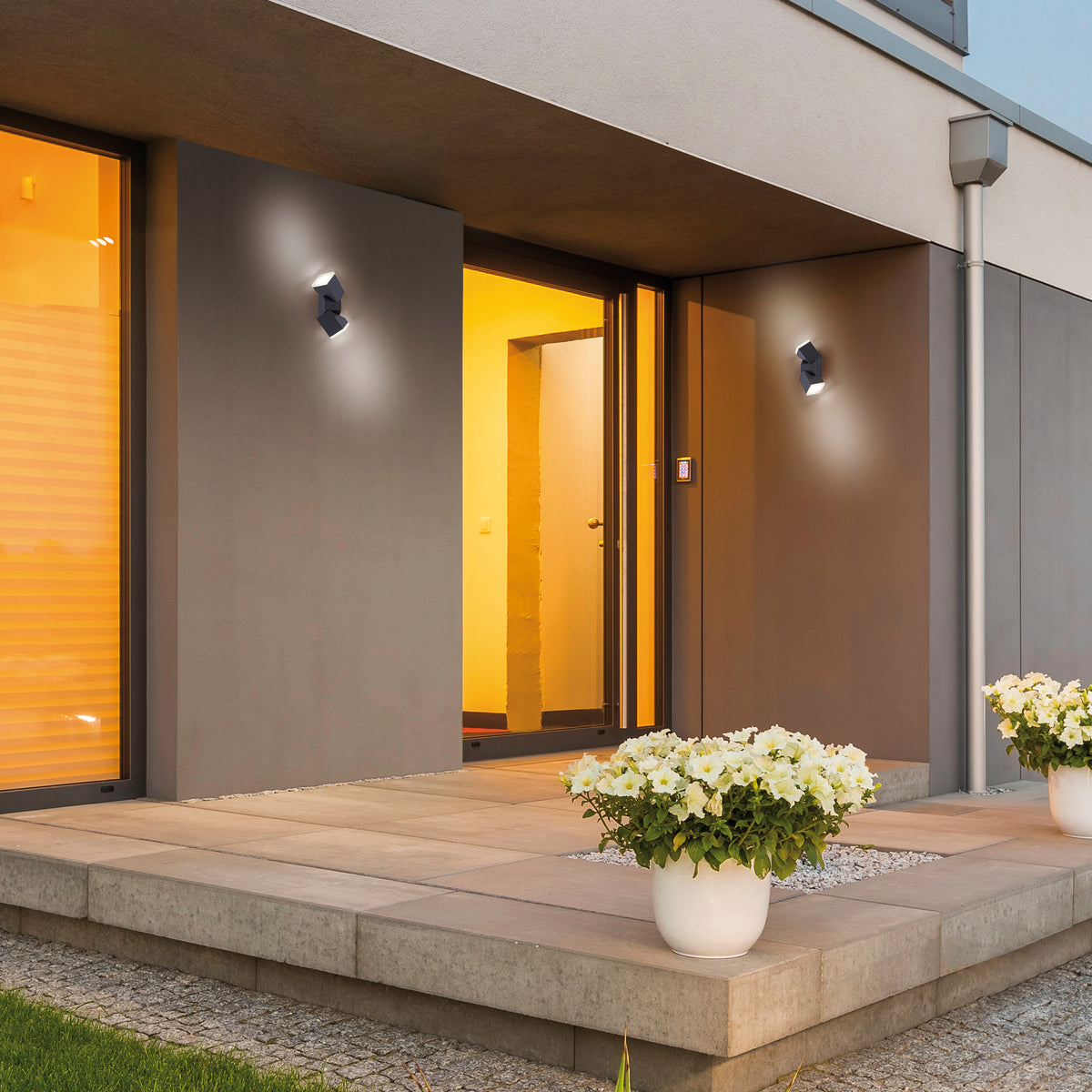 Mise en ambiance extérieure avec deux appliques anthracite éclairant une façade sombre près d'une porte vitrée.