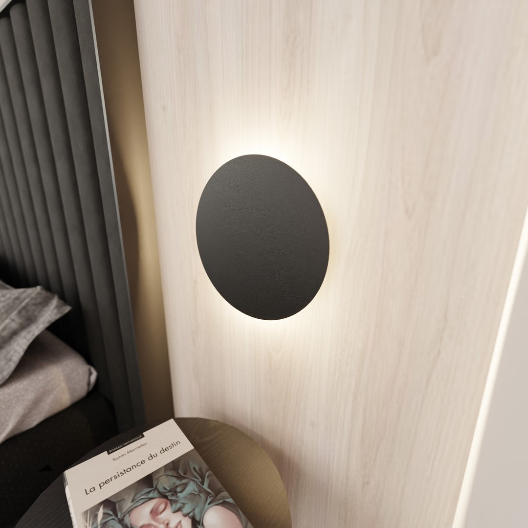 Applique ronde noire allumée sur mur en bois clair, halo lumineux diffus.