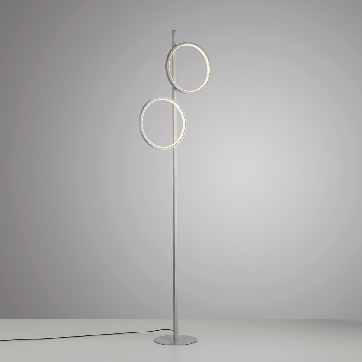 Lampadaire argenté vu de face sur fond gris, câble d'alimentation visible au sol.