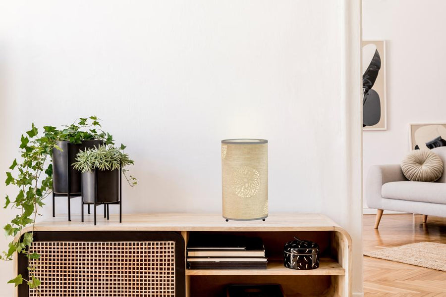 Lampe beige éteinte sur le meuble, entourée de plantes vertes et d'objets noirs.