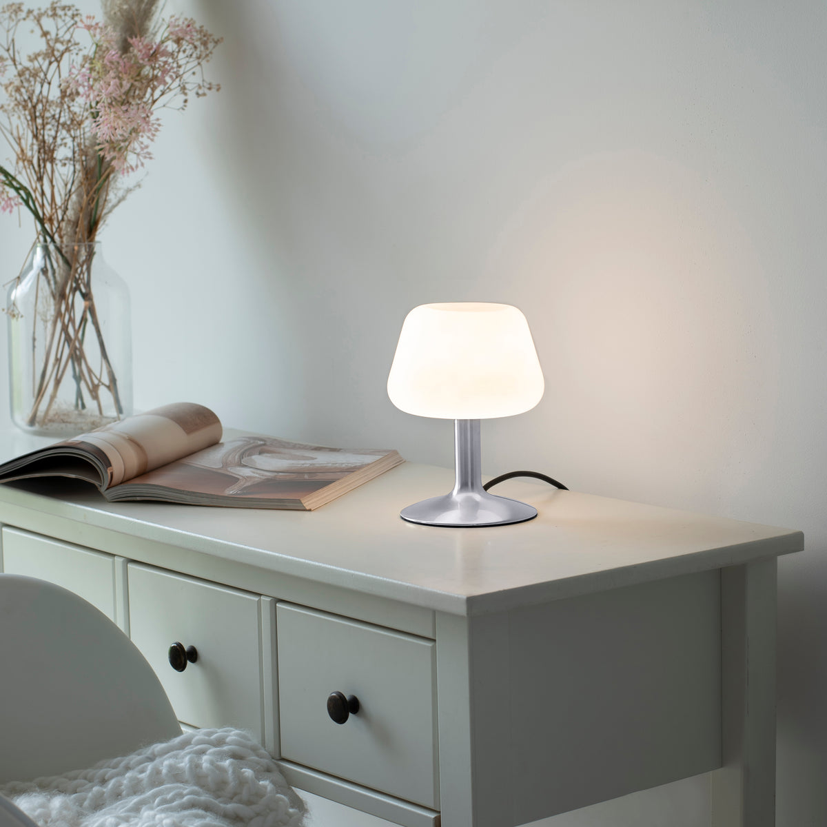 Mise en ambiance sur une commode blanche, lampe allumée près d'un magazine ouvert